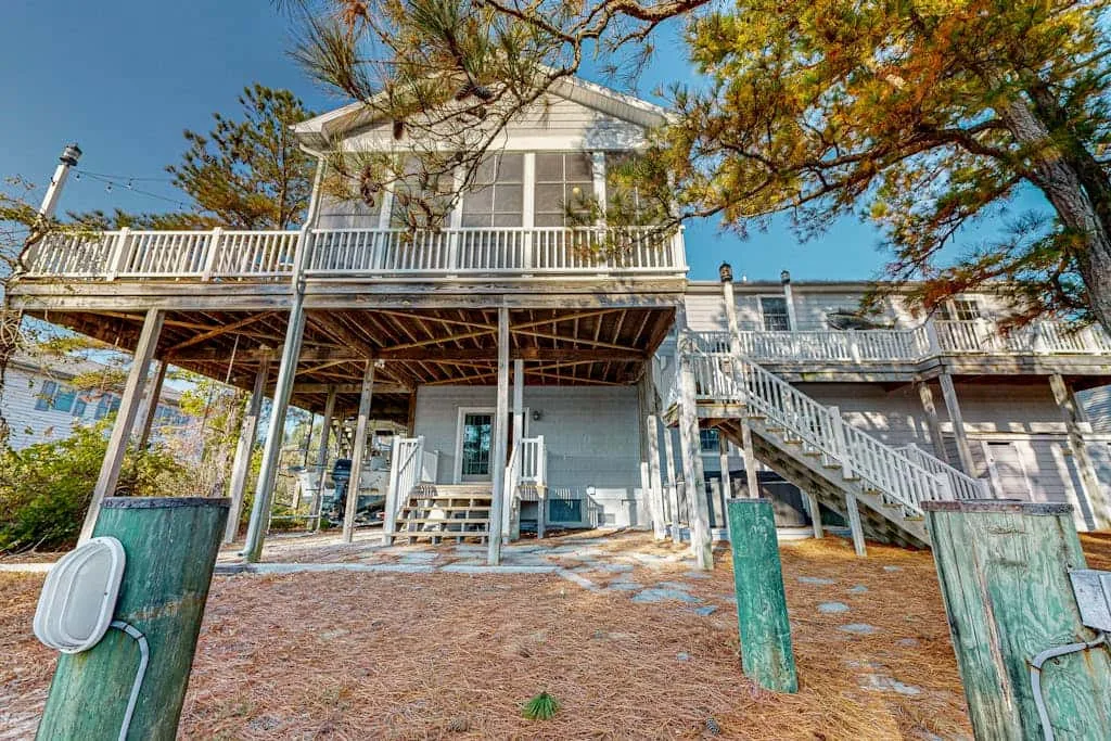 A raised house with wraparound decks and an exterior staircase beneath trees.