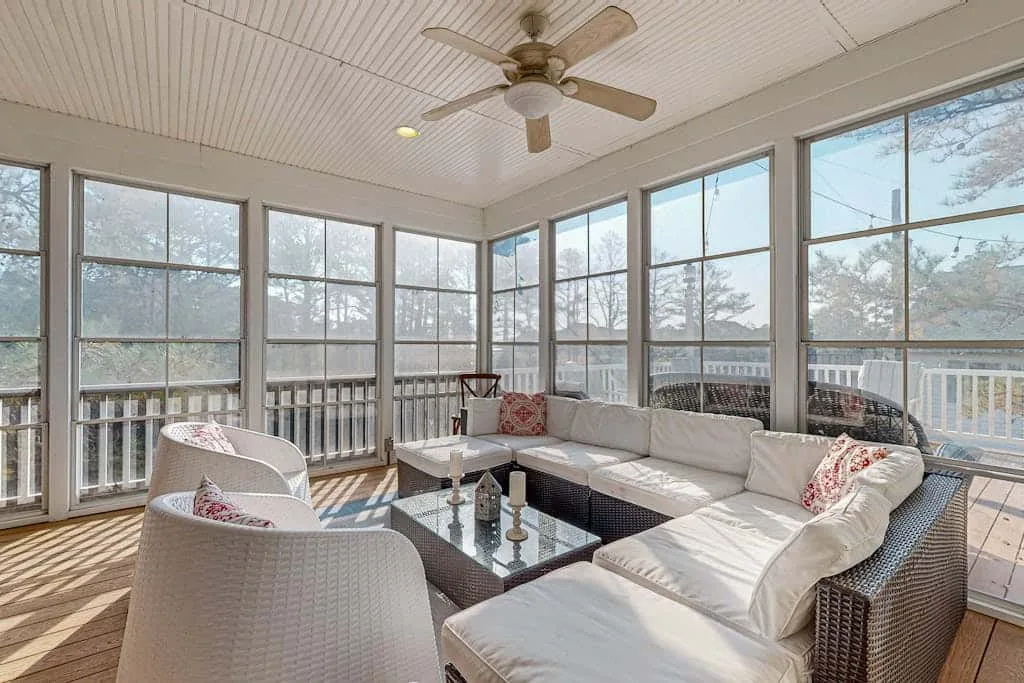 A bright sunroom with cushioned seating, a glass coffee table, and windows overlooking trees.
