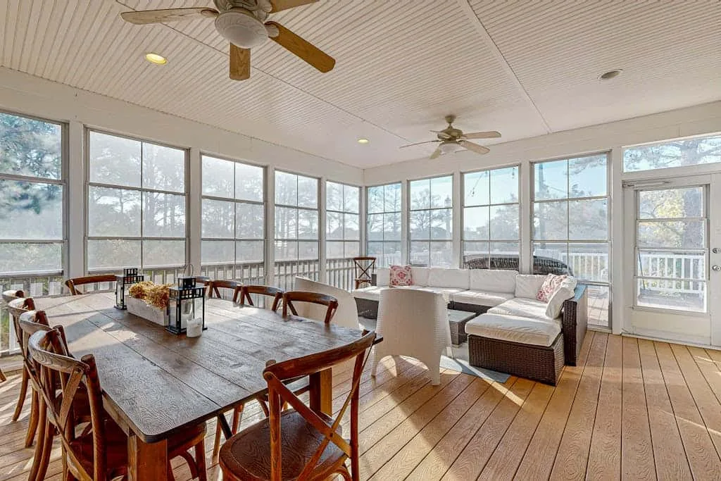 A bright sunroom has a large dining table and seating beside wraparound windows.