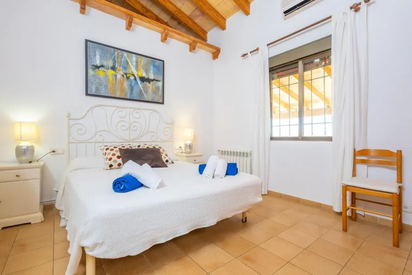 A bright bedroom with a neatly made bed beneath exposed wooden ceiling beams.