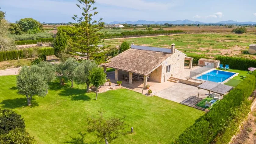 A stone house sits beside a swimming pool in a landscaped rural setting.