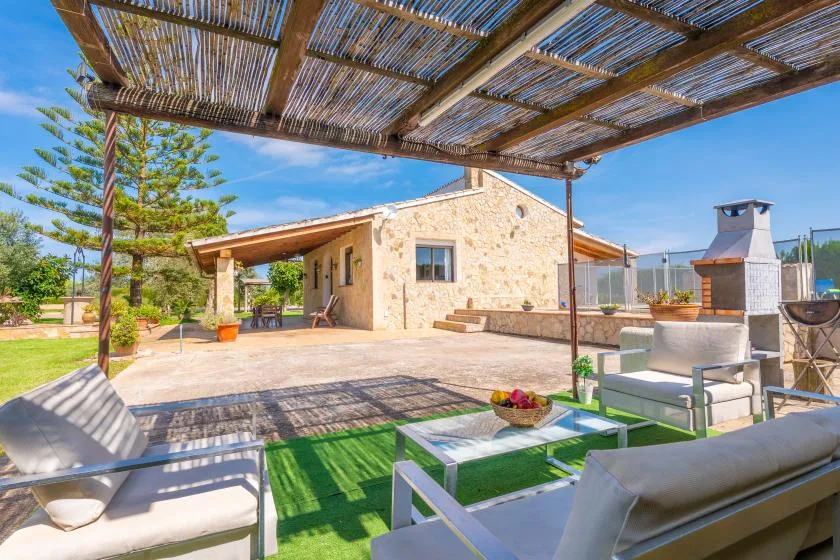A stone house opens onto a sunny patio with outdoor seating beneath a reed pergola.