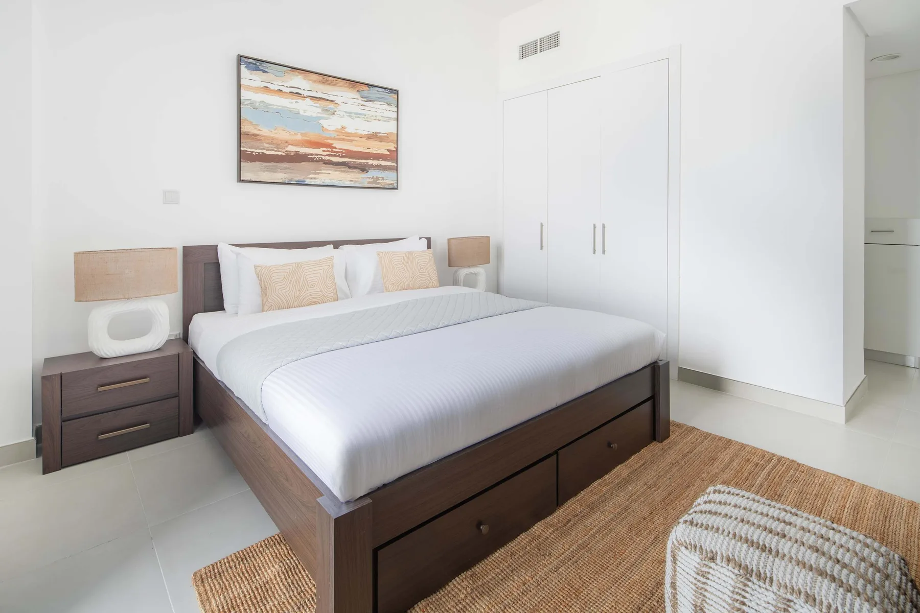 Un dormitorio luminoso con una cama bien tendida y un cuadro enmarcado encima.