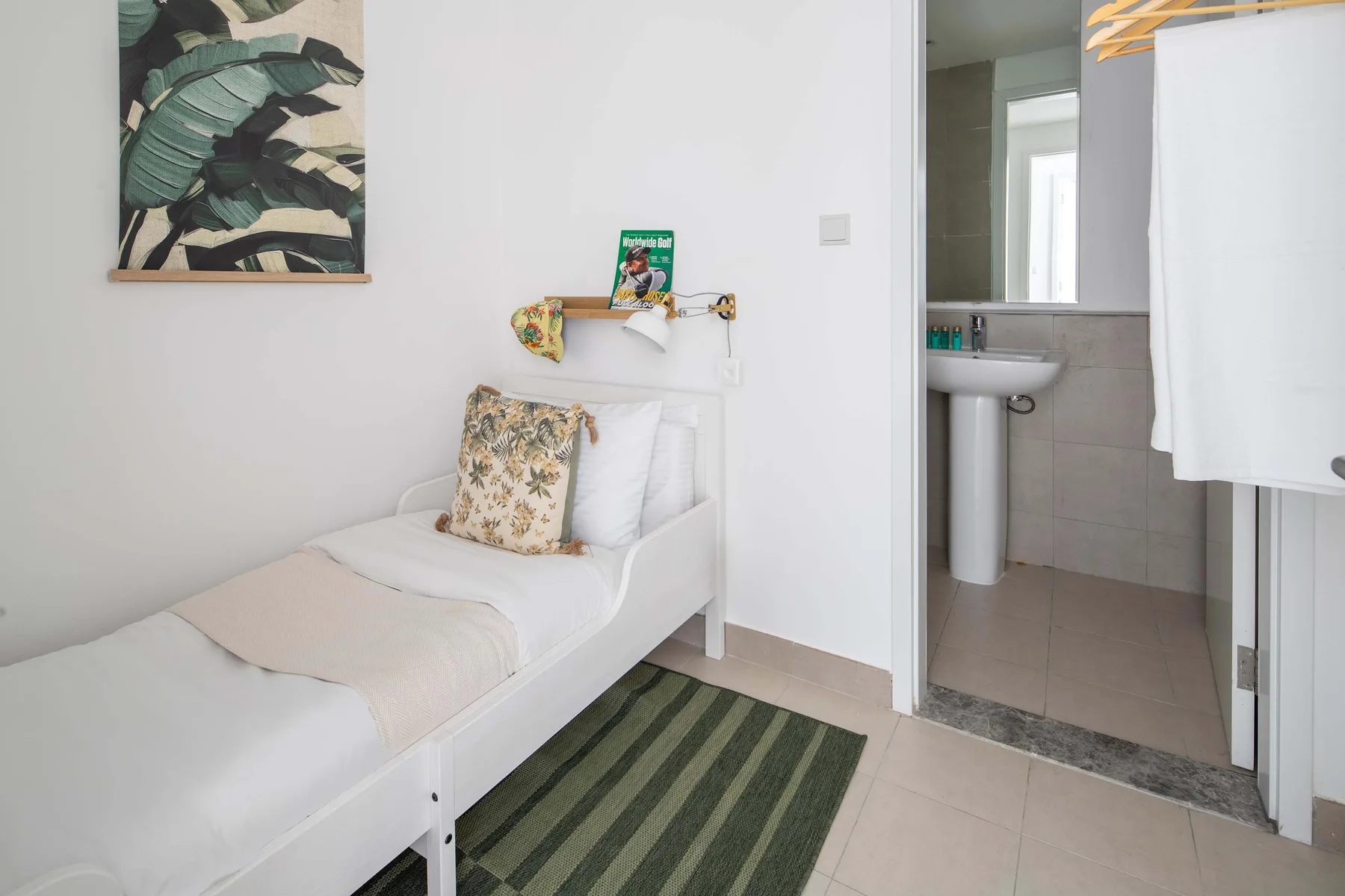Una cama individual está junto a un baño abierto con lavabo y cortina de ducha.