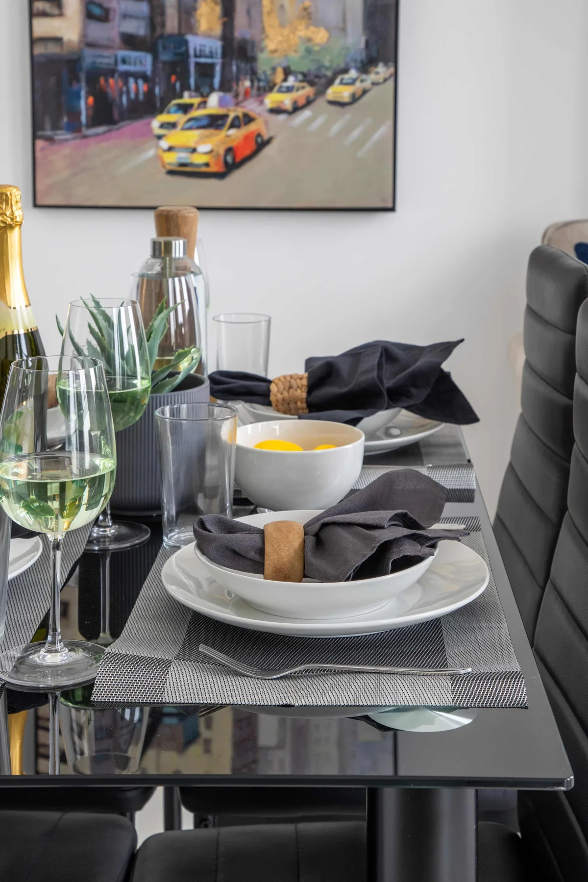 Una mesa de comedor está puesta con platos, vasos y servilletas dobladas bajo una lámina enmarcada de una ciudad.