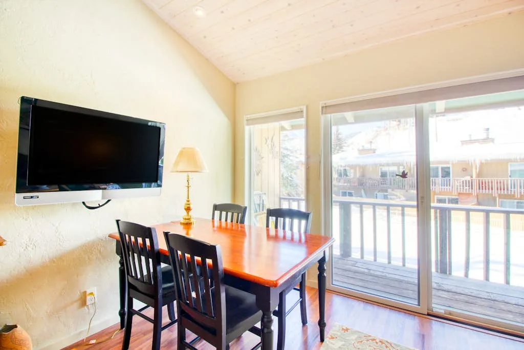A dining area with a wooden table and chairs beside large windows.