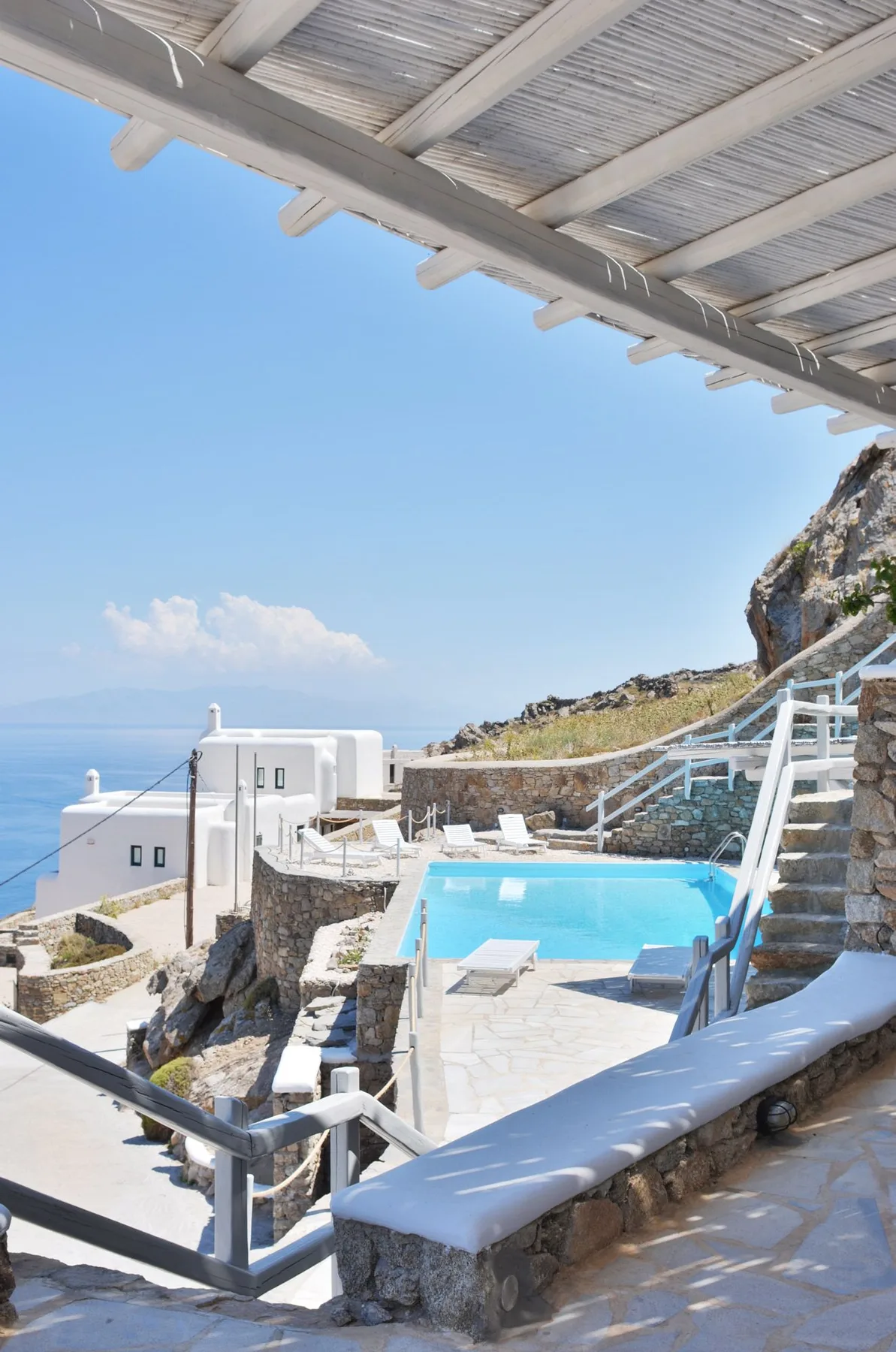 A hillside pool terrace overlooks the sea, with white buildings nearby.