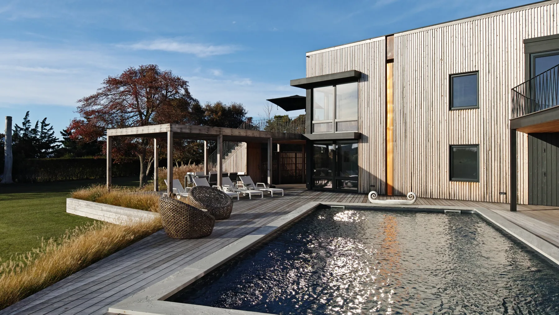 Una casa moderna junto a una piscina, con una terraza de madera y asientos al aire libre.