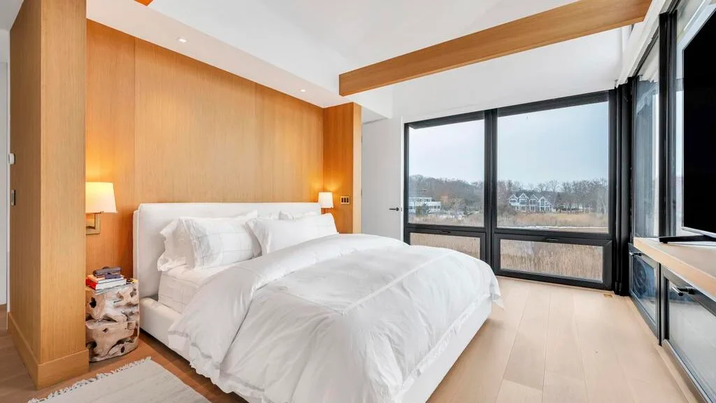 Un dormitorio con una cama blanca junto a grandes ventanas con vistas al exterior.