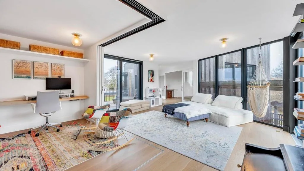 Sala de estar luminosa con zona de asientos, grandes ventanas y un escritorio contiguo.