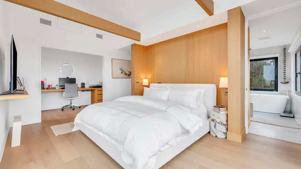 Un dormitorio luminoso con una cama blanca, una pared revestida de madera y un baño contiguo.