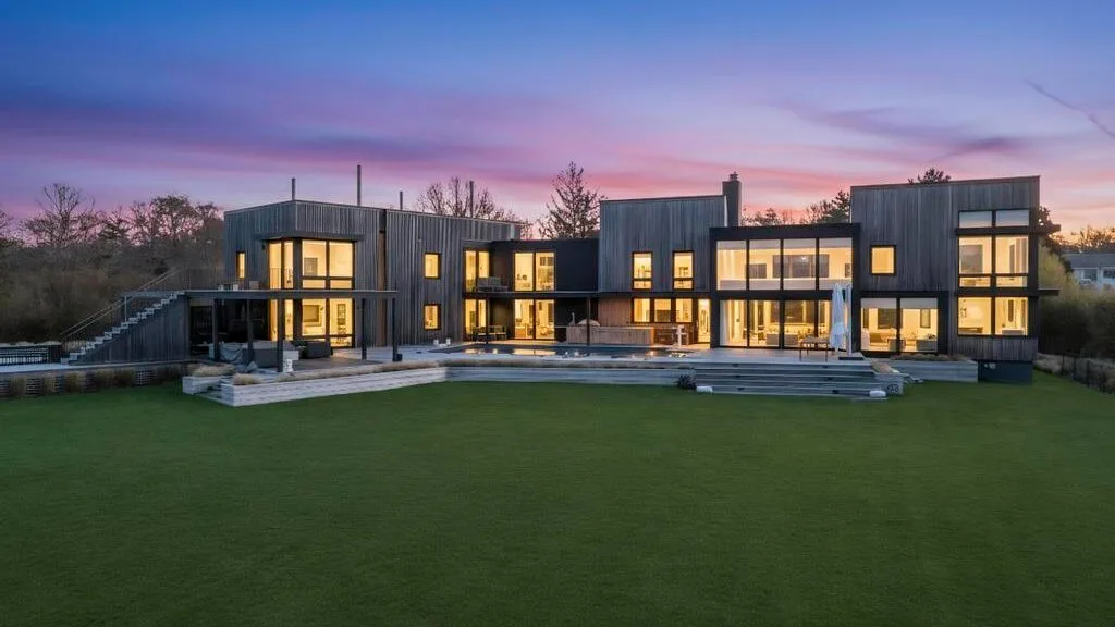 Una casa moderna con ventanas iluminadas se alza junto a un amplio césped al anochecer.
