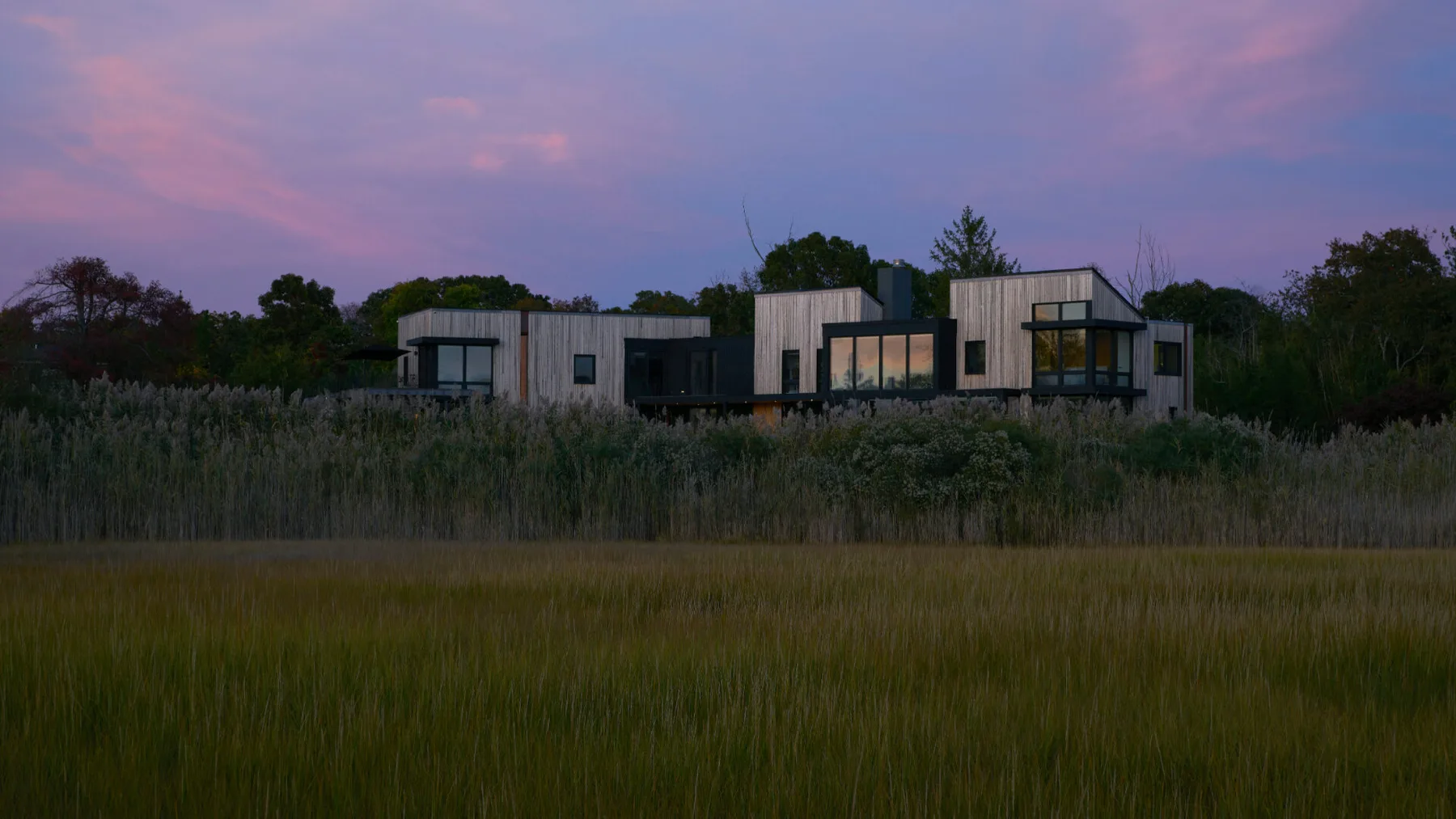 Una casa moderna se encuentra tras un campo de hierba bajo un cielo rosado al atardecer.