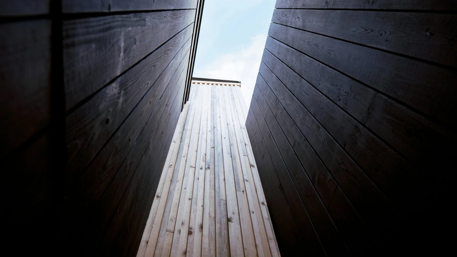 Un pasaje exterior estrecho entre paredes de madera oscura se abre al cielo.