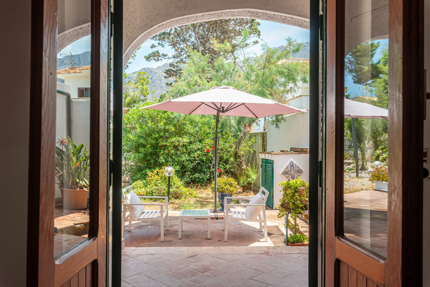 A patio with outdoor seating and a pink umbrella overlooks a green garden.