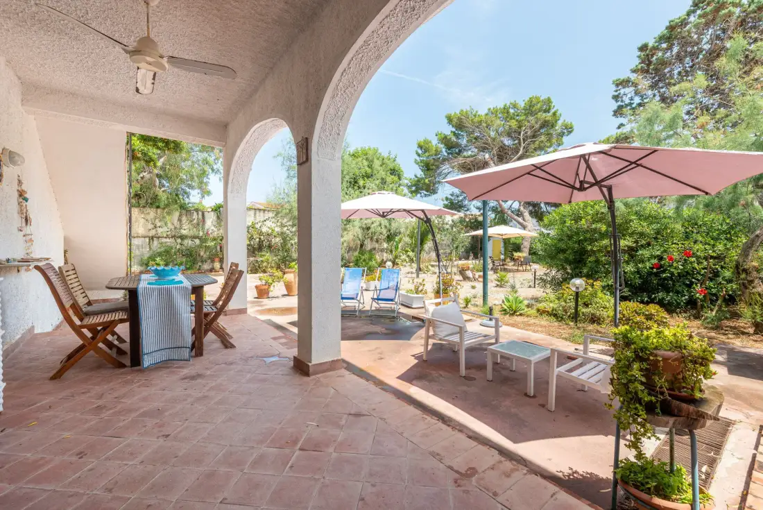 A covered patio opens onto a sunny garden with outdoor seating and umbrellas.