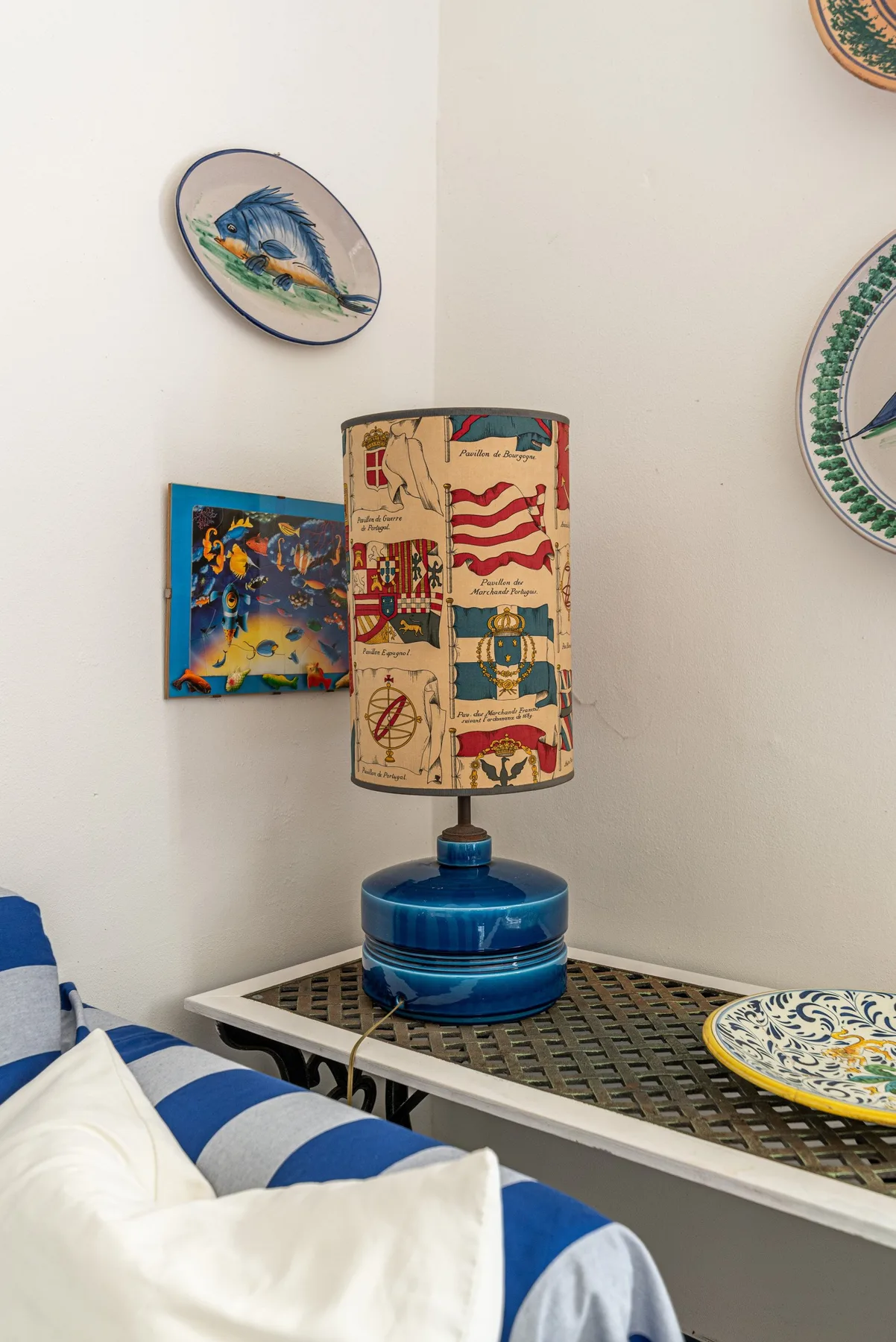 A bedroom corner with a blue-and-white bed, a decorated lamp, and wall plates.