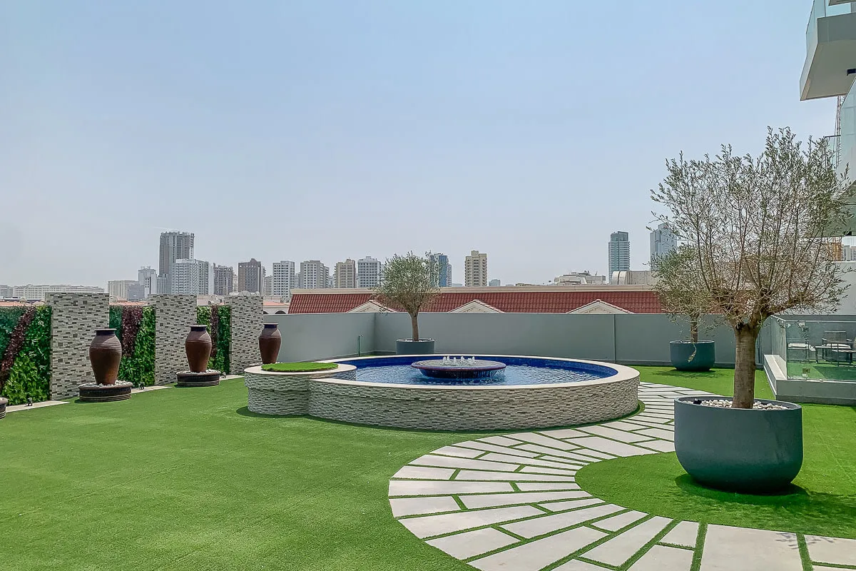 A rooftop terrace with a circular fountain, green lawn, and city skyline.
