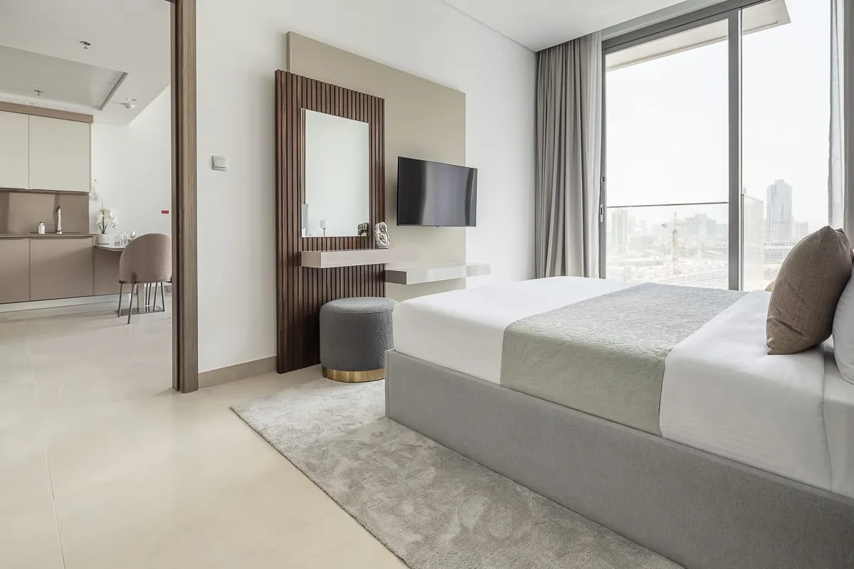 A bright bedroom with a large bed, a wall-mounted television, and floor-to-ceiling windows.