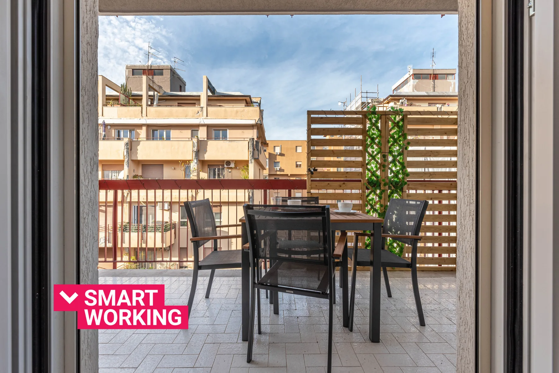 A furnished balcony with a table and seating overlooks nearby buildings.