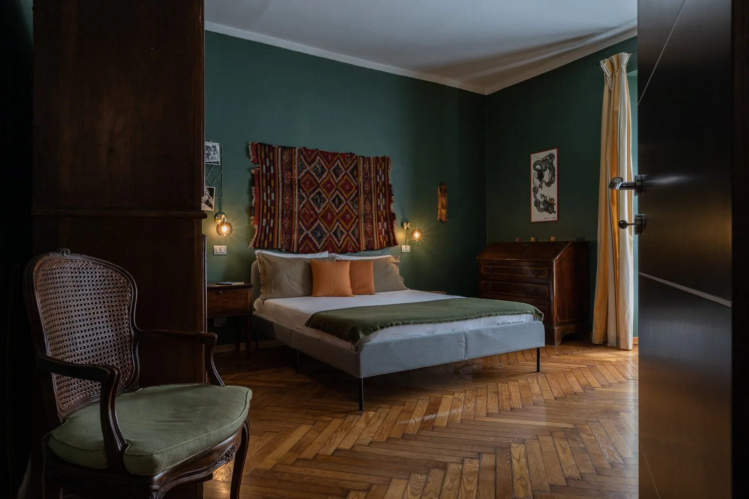 Un dormitorio con una cama, paredes verde oscuro y un tejido estampado sobre el cabecero.