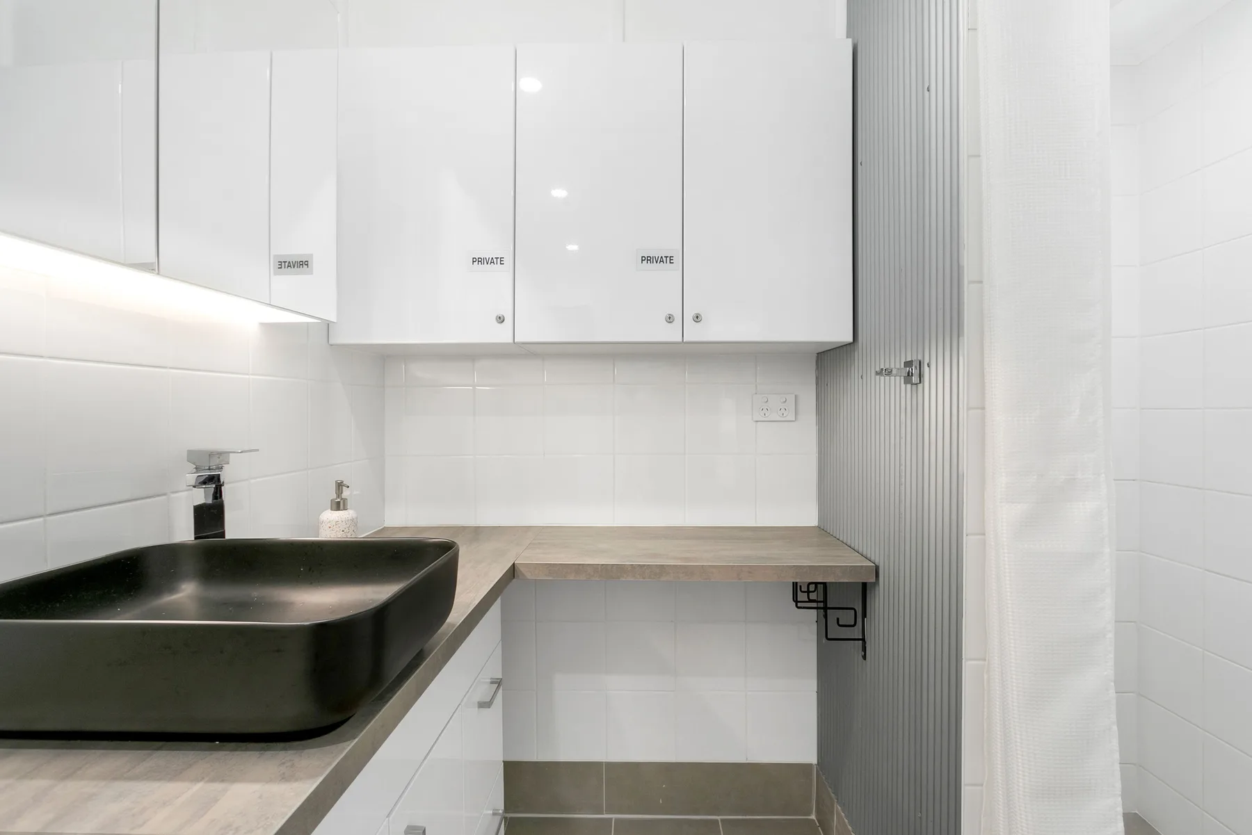 Un baño luminoso con un lavabo negro, armarios blancos y una cortina de ducha.