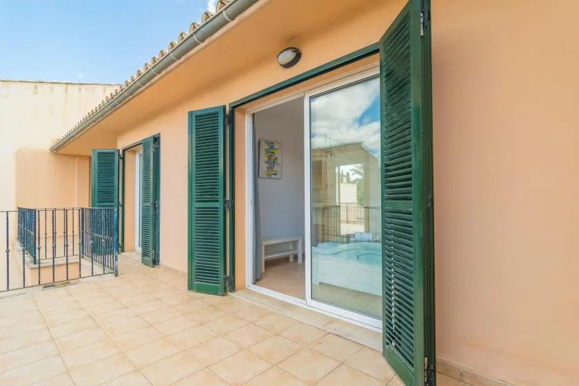 Una terraza de baldosas bordea un edificio color durazno con contraventanas verdes abiertas.