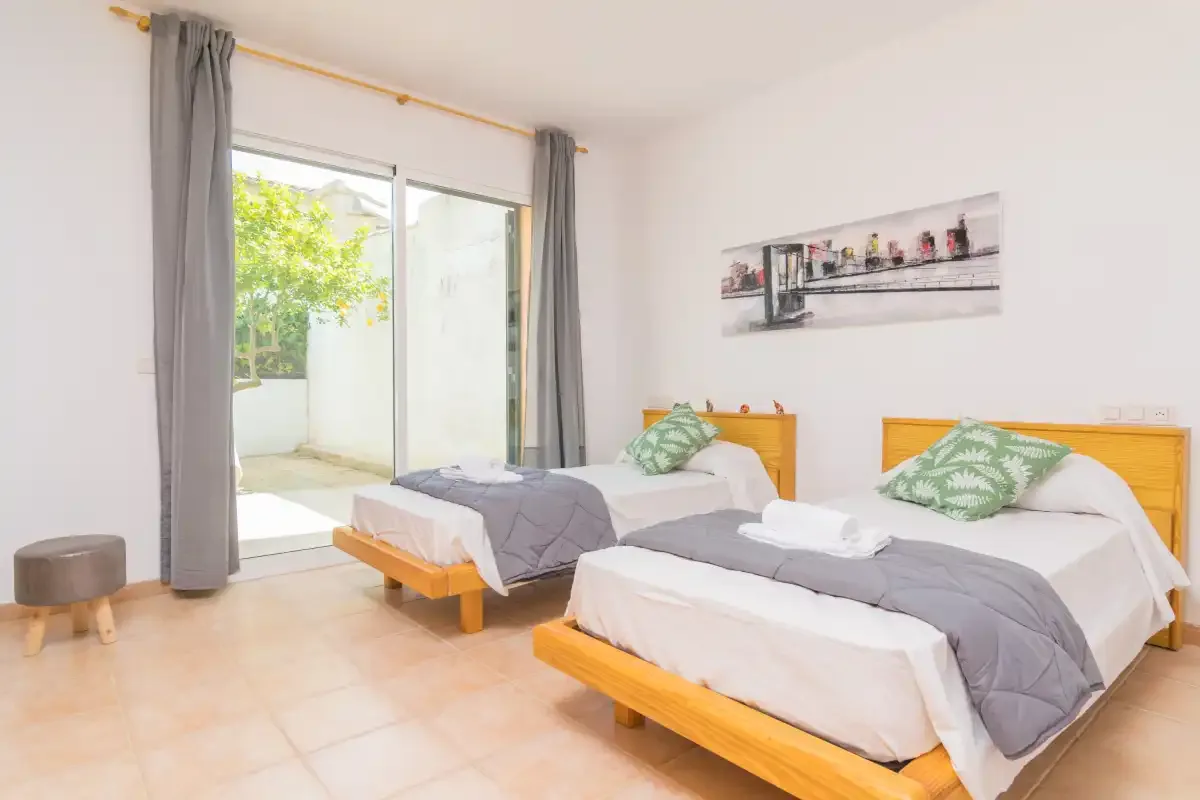 Un dormitorio luminoso con dos camas, ropa de cama gris y una puerta de cristal que da a un patio.