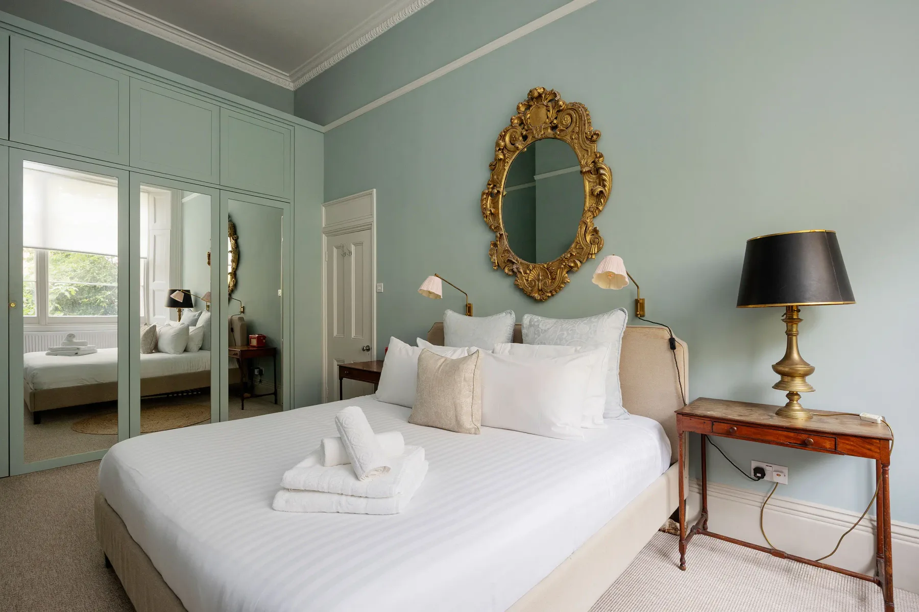 Dormitorio con una cama bien tendida, armarios con puertas espejadas y un espejo ornamental en la pared.