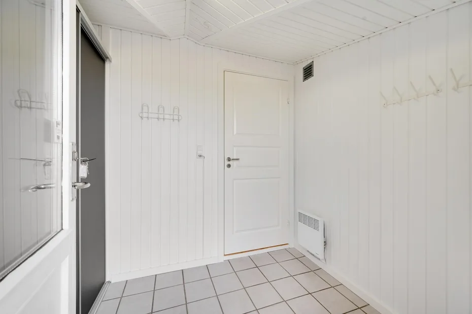 Una pequeña entrada blanca con suelo de baldosas, ganchos en la pared y una puerta cerrada.