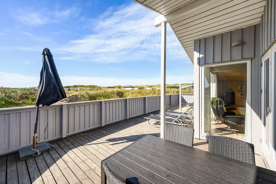 Una terraza de madera con asientos al aire libre da a dunas cubiertas de hierba bajo un techo.