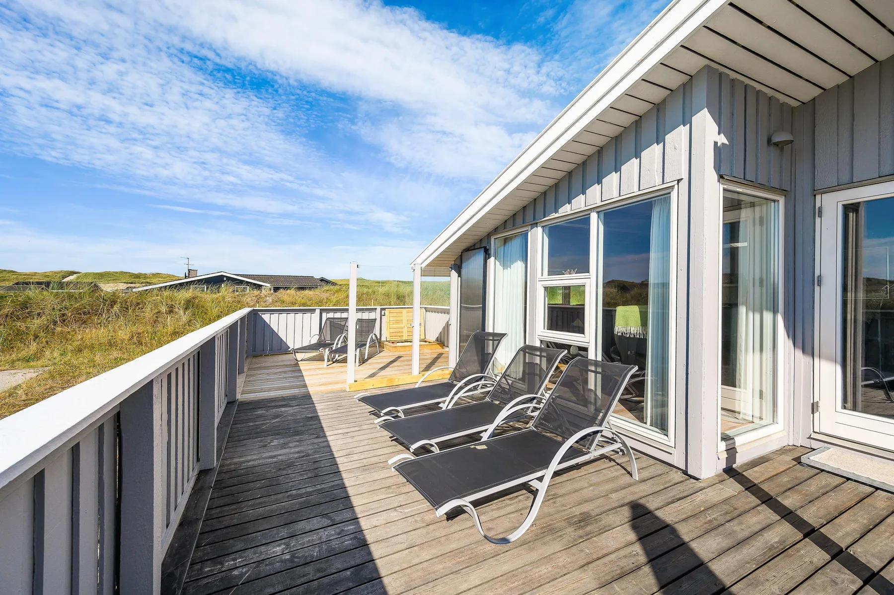 Una terraza de madera soleada junto a una casa, con asientos exteriores y vistas a dunas cubiertas de hierba.
