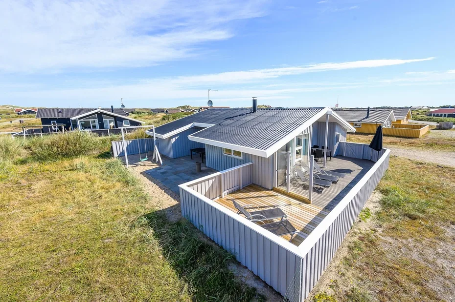 Una casa moderna con una terraza de madera cercada se encuentra entre dunas cubiertas de hierba.