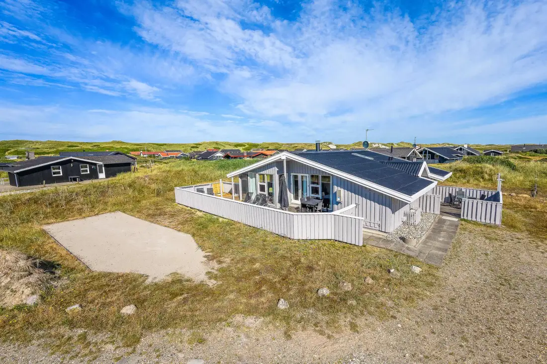 Una casa gris con una terraza blanca está entre dunas cubiertas de hierba bajo un cielo azul.