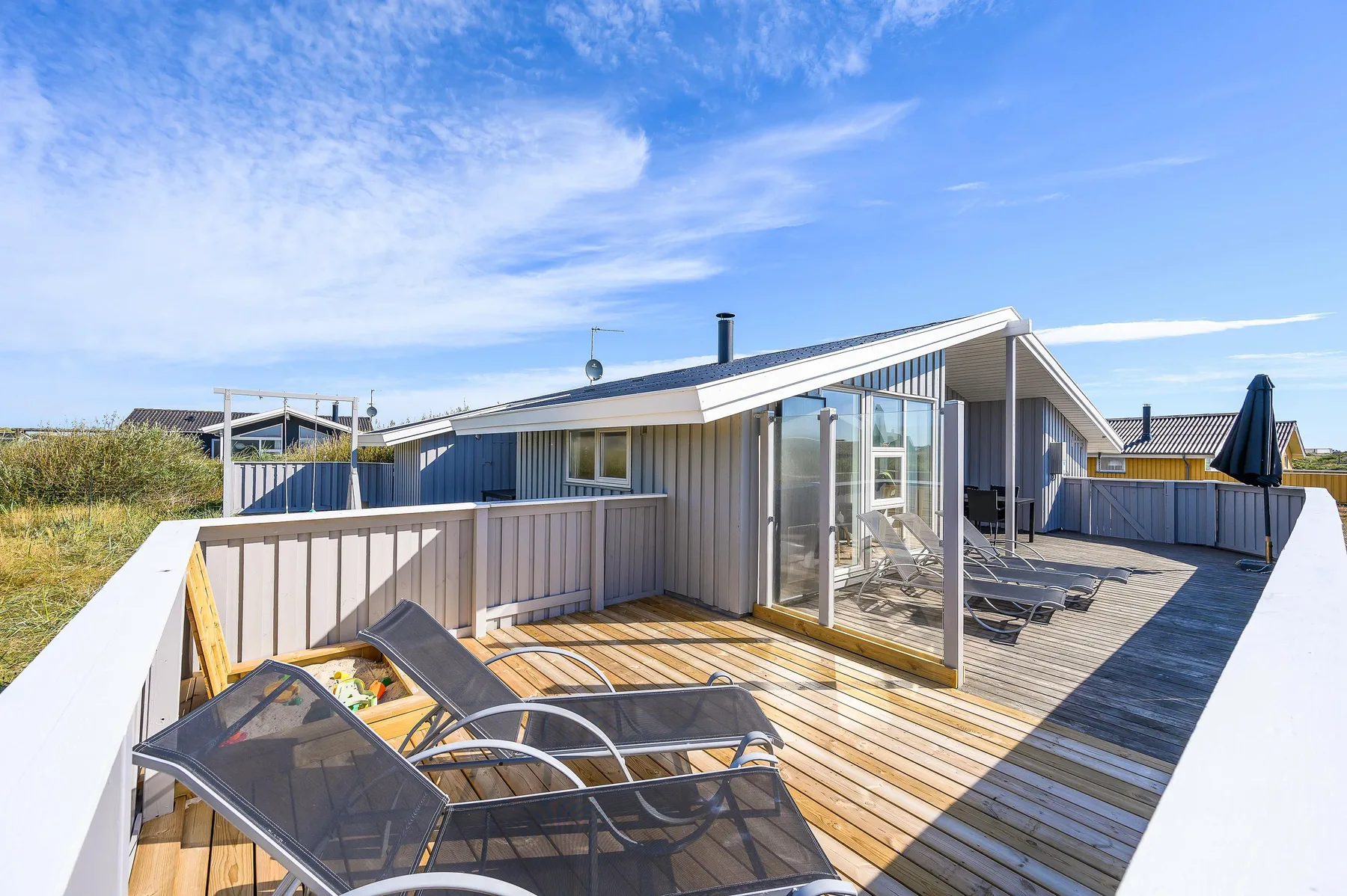 Una terraza de madera soleada junto a una casa moderna, con asientos reclinables y puertas de cristal.