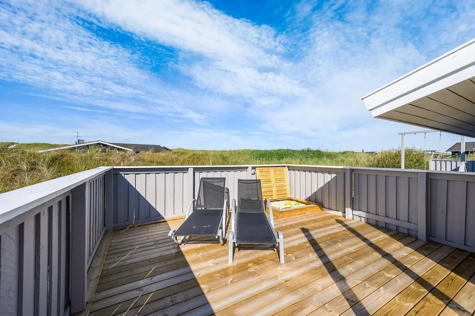 Una terraza de madera con tumbonas reclinables da a dunas cubiertas de hierba bajo un cielo azul.