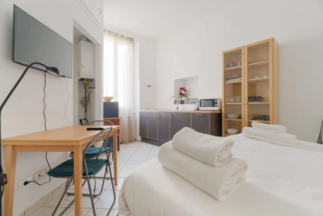 A bright room with a bed, a desk, and a kitchenette along the back wall.