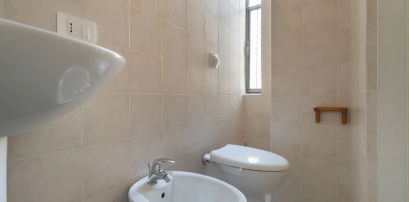 A tiled bathroom with a toilet, sink, and narrow window.
