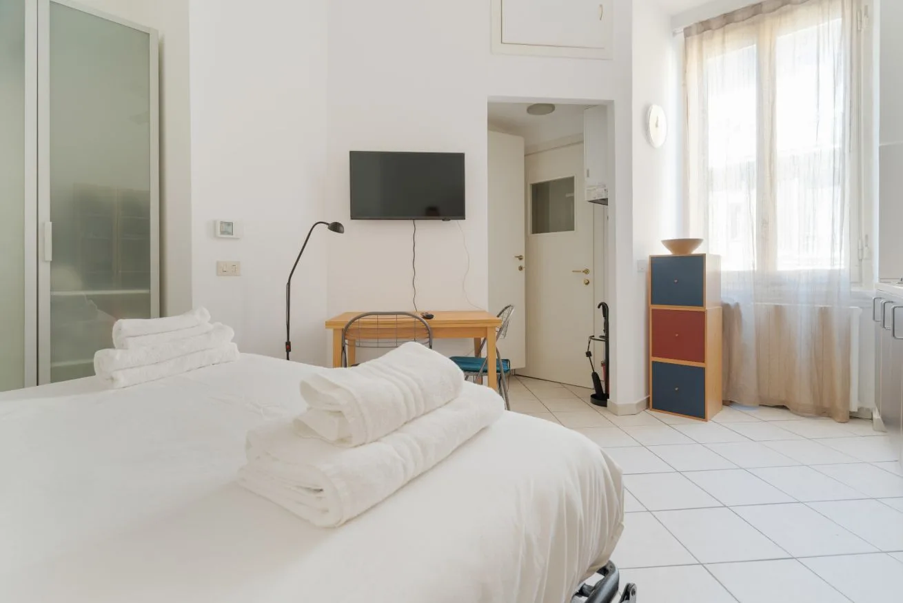 A bright bedroom with white bedding, a television, and a sunlit window.