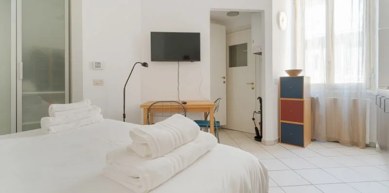 A bright bedroom with white bedding, a wall-mounted television, and a large window.