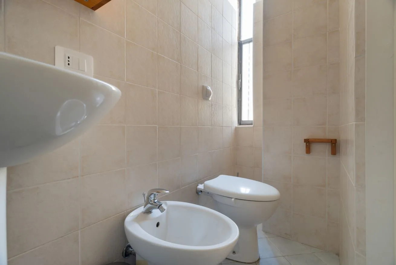 A tiled bathroom with a sink, toilet, and narrow window.