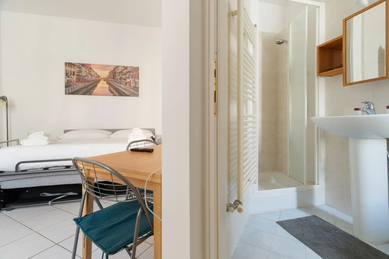 A bedroom with a bed and desk is visible beside a bathroom with a shower and sink.