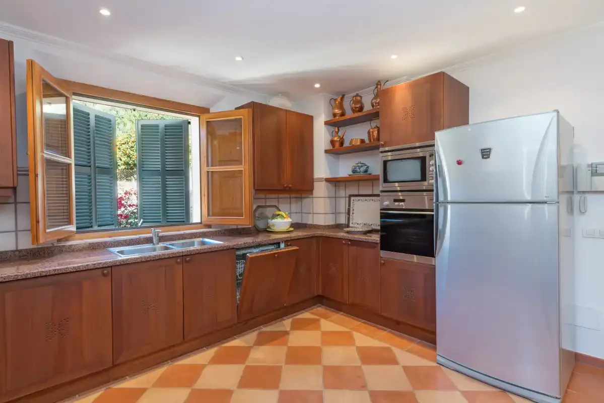 A kitchen with wooden cabinets, a refrigerator, and a window overlooking greenery.