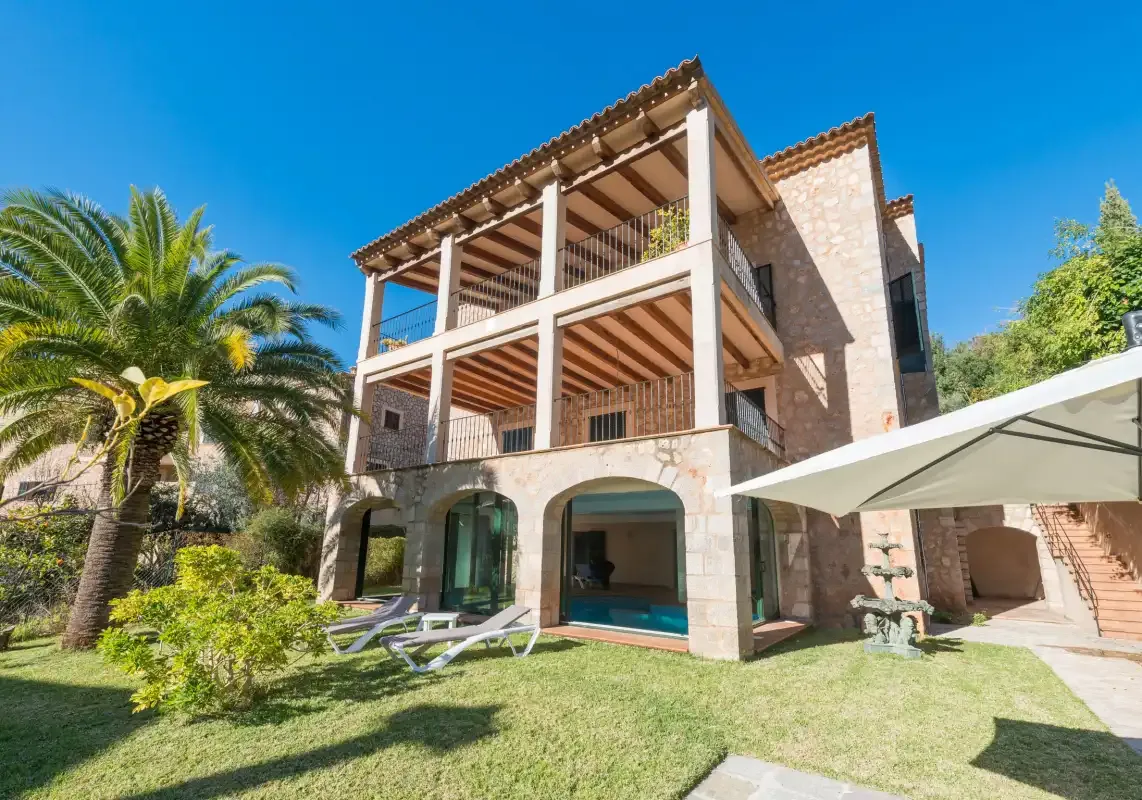 A stone house with a covered terrace overlooks a garden and swimming pool.