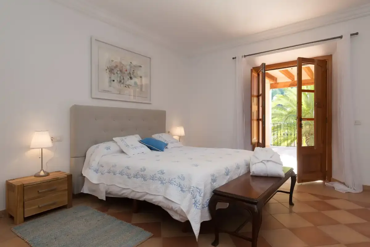 A bright bedroom with a made bed and open wooden doors to a balcony.