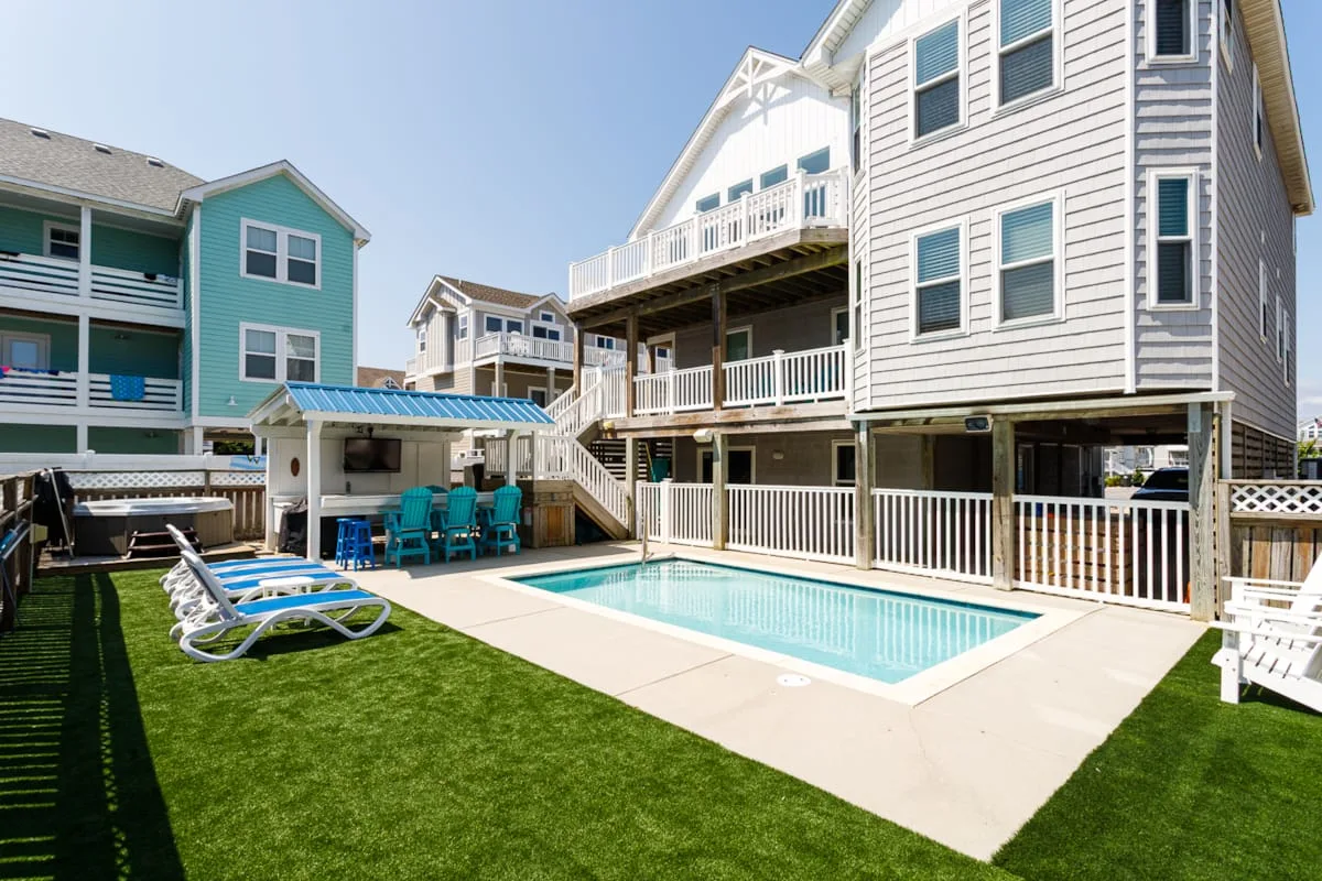 A backyard swimming pool sits beside a large coastal-style house and lawn.