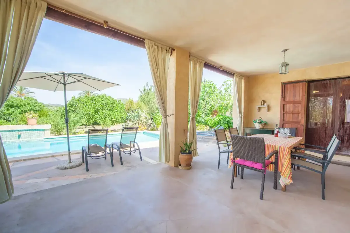 A covered patio with outdoor dining furniture overlooks a swimming pool and garden.