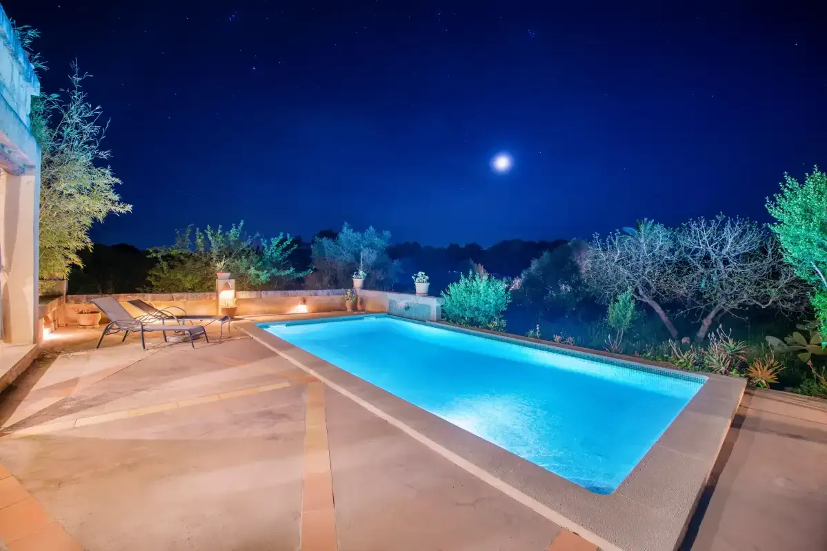 An outdoor swimming pool glows beneath the night sky, with seating along the paved deck.