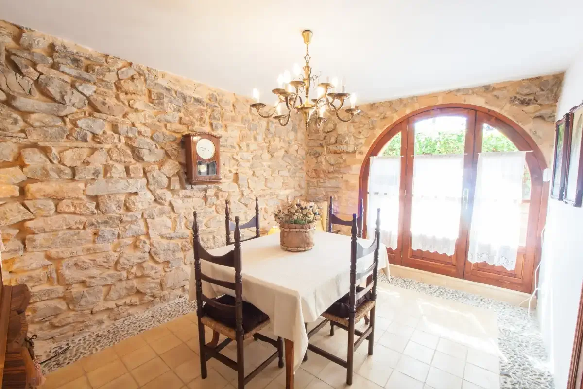 A rustic dining room has a table set beneath a chandelier beside arched glass doors.