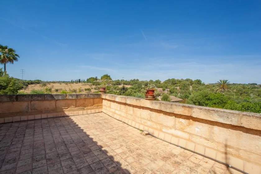 A sunlit tiled terrace overlooks a green landscape beneath a clear blue sky.